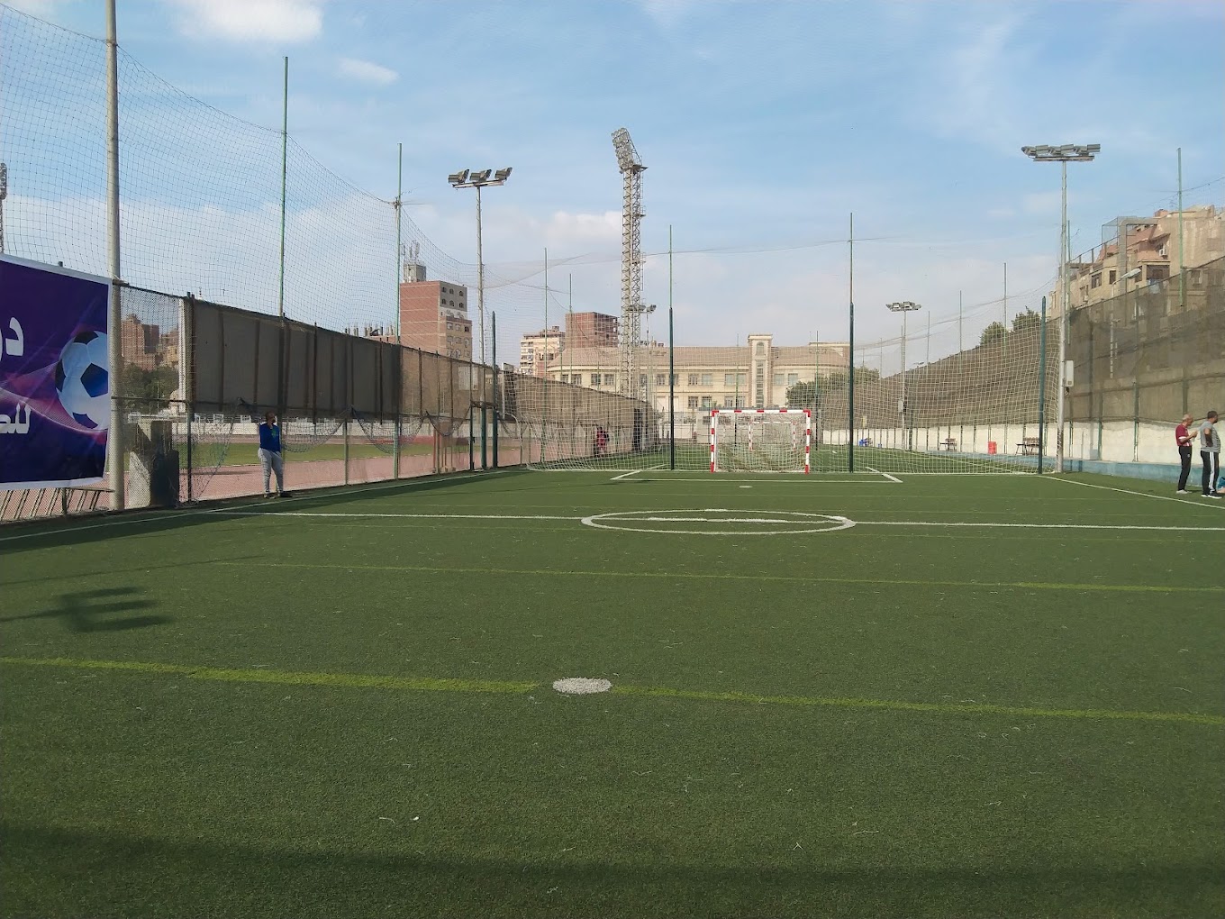 Eingezäunter Kunstrasen-Mini-Fußballplatz mit Tor und Flutlichtmasten.