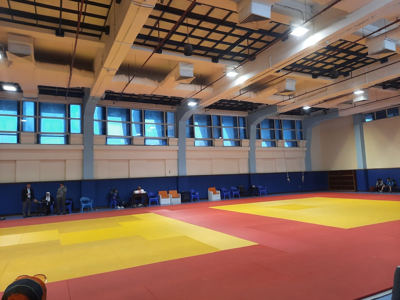 Indoor-Kampfsporthalle mit großer Mattenfläche und Sitzgelegenheiten entlang der Wand.