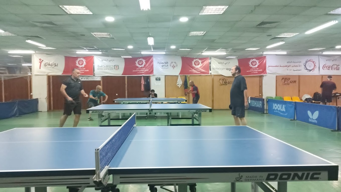 Tischtennishalle mit mehreren Tischen und trainierenden Spielern hinter Absperrungen.