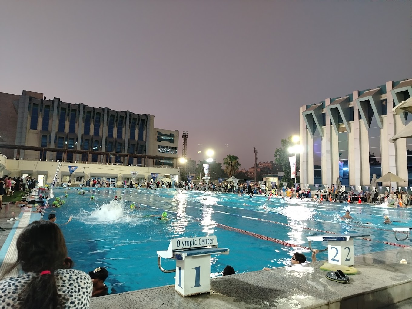 Belebte Abendsession im Outdoor-Becken unter Flutlicht, Schwimmer in den Bahnen und Personen am Beckenrand.