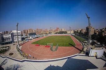 Aerial view of red running track around green field, empty stands and city skyline under blue sky, training camp venue