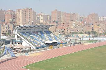 Roofed grandstand with blue and yellow seats beside a red running track and green field; urban backdrop, training camp