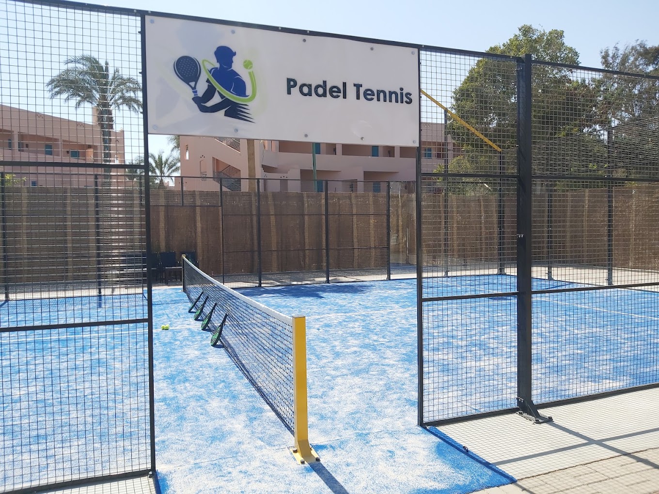 Entrée du court de padel tennis avec filet, clôture et panneau 'Padel Tennis'.
