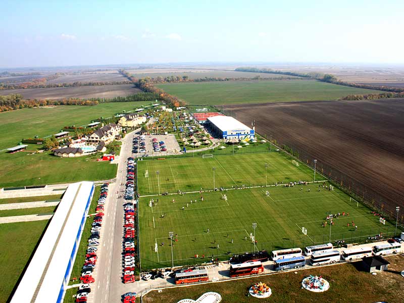Luftaufnahme der Anlage mit Sportplätzen, Gebäuden, Parkflächen und umliegender Landschaft.
