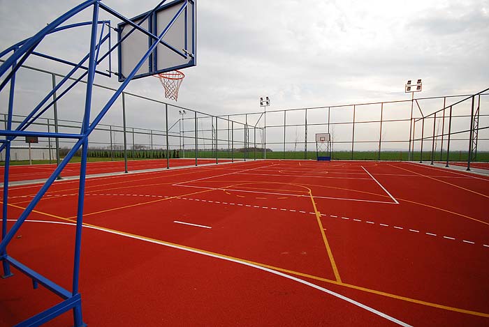 Outdoor-Multisportplatz mit Basketballkörben und markierten Linien auf rotem Belag, umzäunt.