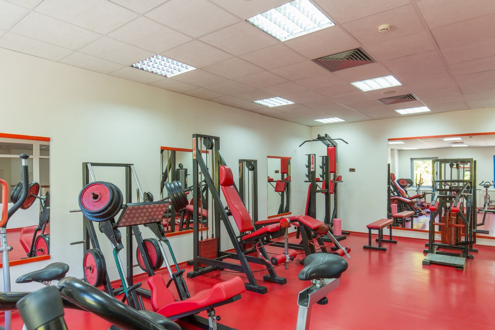 Innen-Fitnessraum mit Kraftgeräten, Bänken und Spiegeln auf rotem Boden.