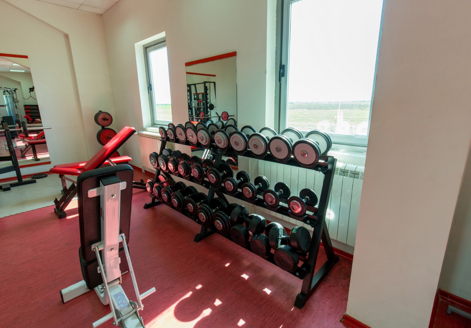 Fitnessraum-Ecke mit Hantelablage, verstellbarer Bank und Spiegeln an großen Fenstern.