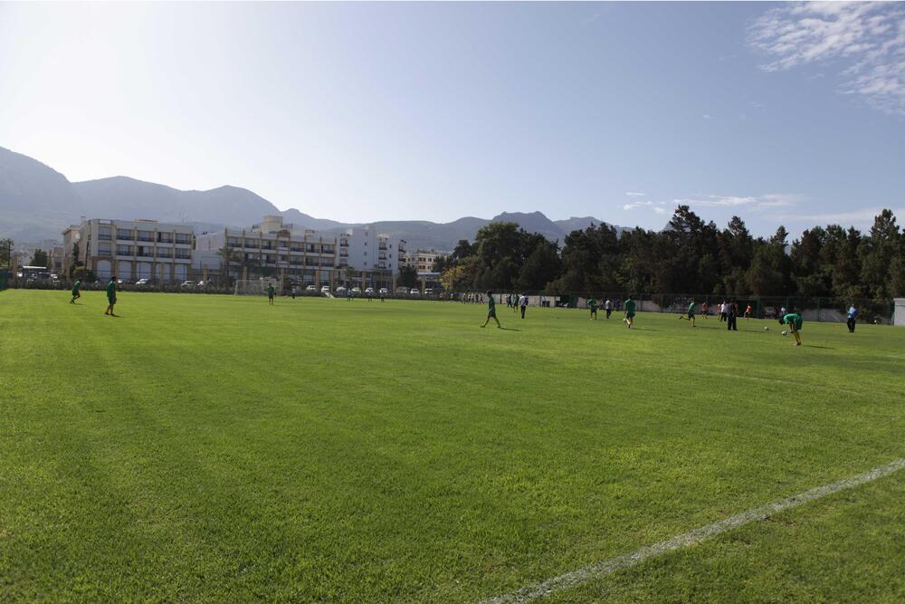 Wide view of a natural-grass football pitch with players training and mountains in the background.