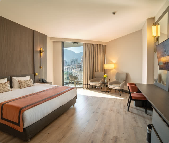 Modern hotel room with a double bed, seating area, work desk and balcony view over the city and mountains.