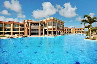 Large resort swimming pool in front of the hotel buildings under a blue sky.