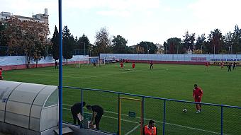 Players in red run football drills on a grass pitch behind a fence, with covered bench on the sideline.