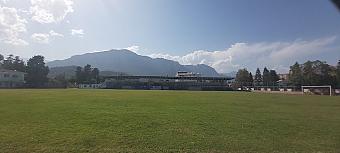 Empty grass football pitch with goalposts, grandstand and mountains under a clear sky.