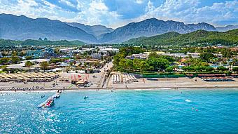 Aerial view of beachfront with sun loungers, pier, and mountains behind the resort.