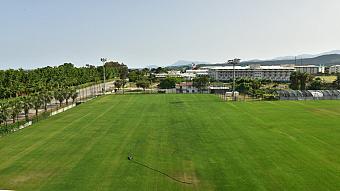 Full-size grass football field with floodlights near resort buildings, viewed from the end line.