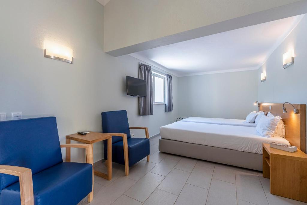 Chambre twin lumineuse avec deux lits simples, TV murale et coin salon avec fauteuils bleus.