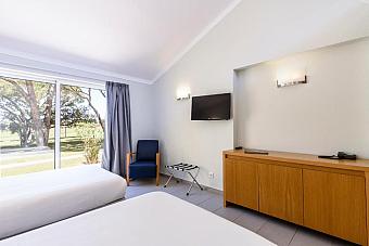 Zweibettzimmer mit TV, Sideboard und großem Fenster zum Gelände.