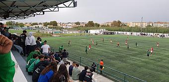 Zuschauer verfolgen ein Fußballspiel auf einem Kunstrasenplatz von einer überdachten Tribüne aus.