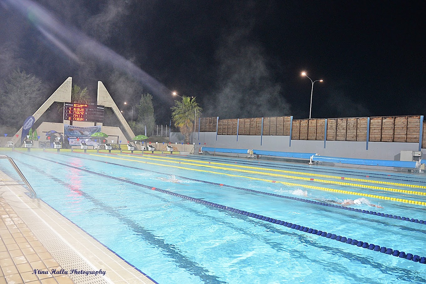 Nächtlicher Schwimmwettkampf in einem beleuchteten Außenbecken mit Anzeigetafel und Bahnmarkierungen.