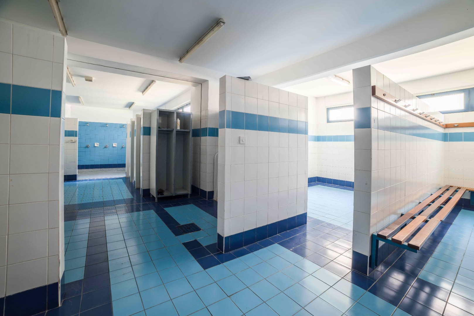 Blau gefliester Umkleideraum mit Bänken, Spinden und Duschbereich.