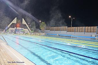 Nighttime swimming race in an illuminated outdoor pool with scoreboard and lane markers.