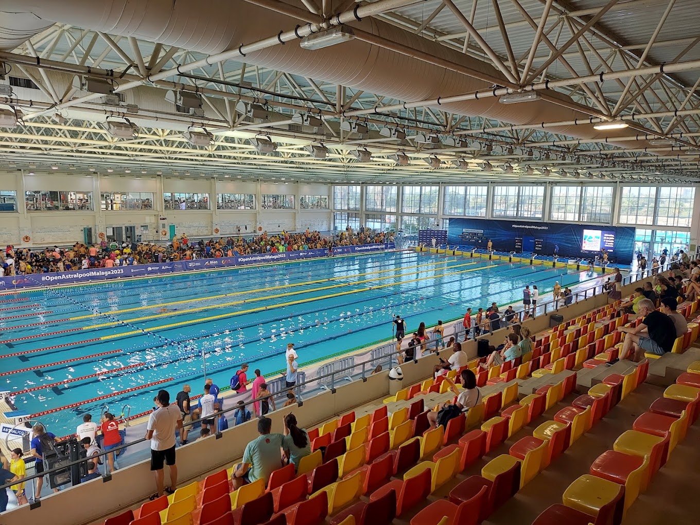 Indoor competition pool during a swim event, viewed from the spectator stands.