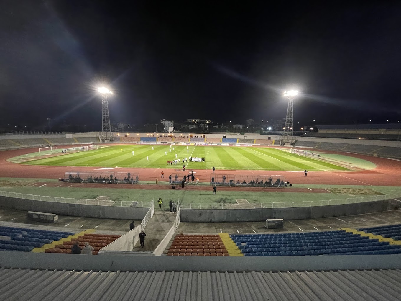Night view of a football match in a floodlit stadium with a surrounding running track.