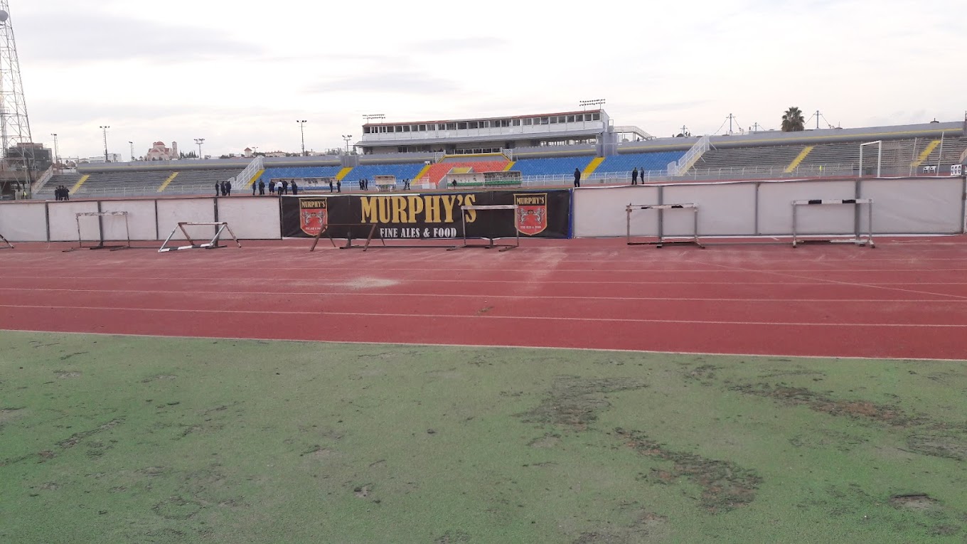 Trackside view of a stadium with seating, floodlight towers and pitch-side barriers.