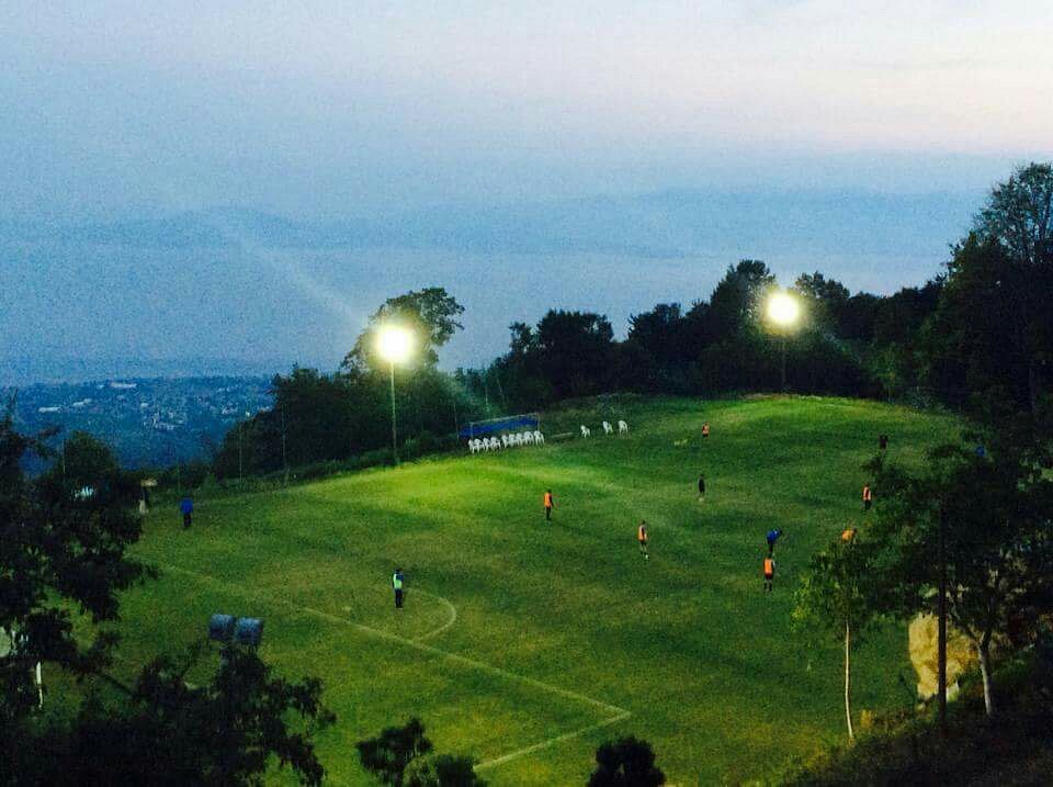 Floodlit football training pitch on a hillside with players at dusk.