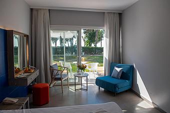 Helles Hotelzimmer mit blauen und grauen Möbeln, Terrassenblick auf einen grünen Garten und kleinem Sitzbereich im Freien