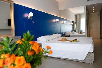 Helles Hotelzimmer mit Doppelbett, blauer Akzentwand und einem Blumenstrauß aus orangefarbenen Blüten