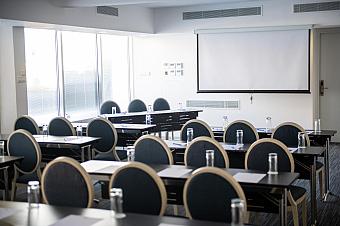 Konferenzraum mit Reihen von Stühlen und Tischen, die auf eine Projektionsleinwand ausgerichtet sind, vorbereitet für ein Coaching oder Teammeeting