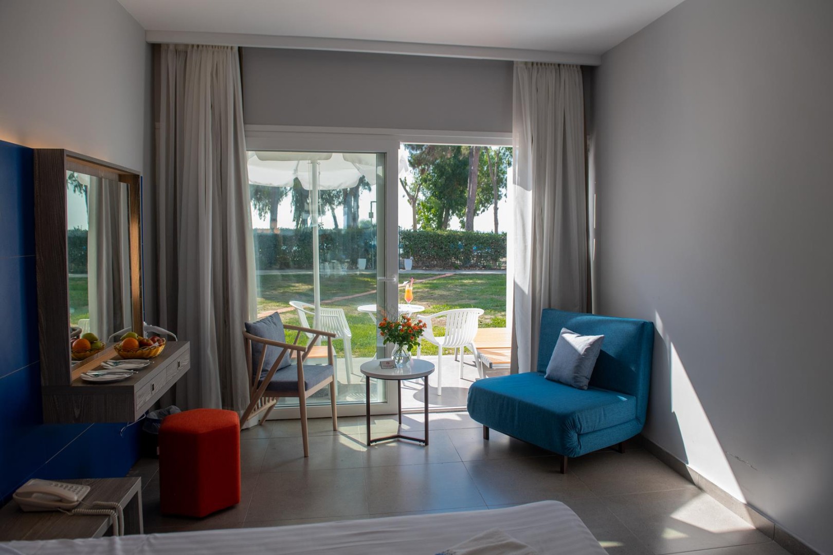 Bright hotel room with blue and gray furniture, a terrace view of a green garden, and a small outdoor seating area