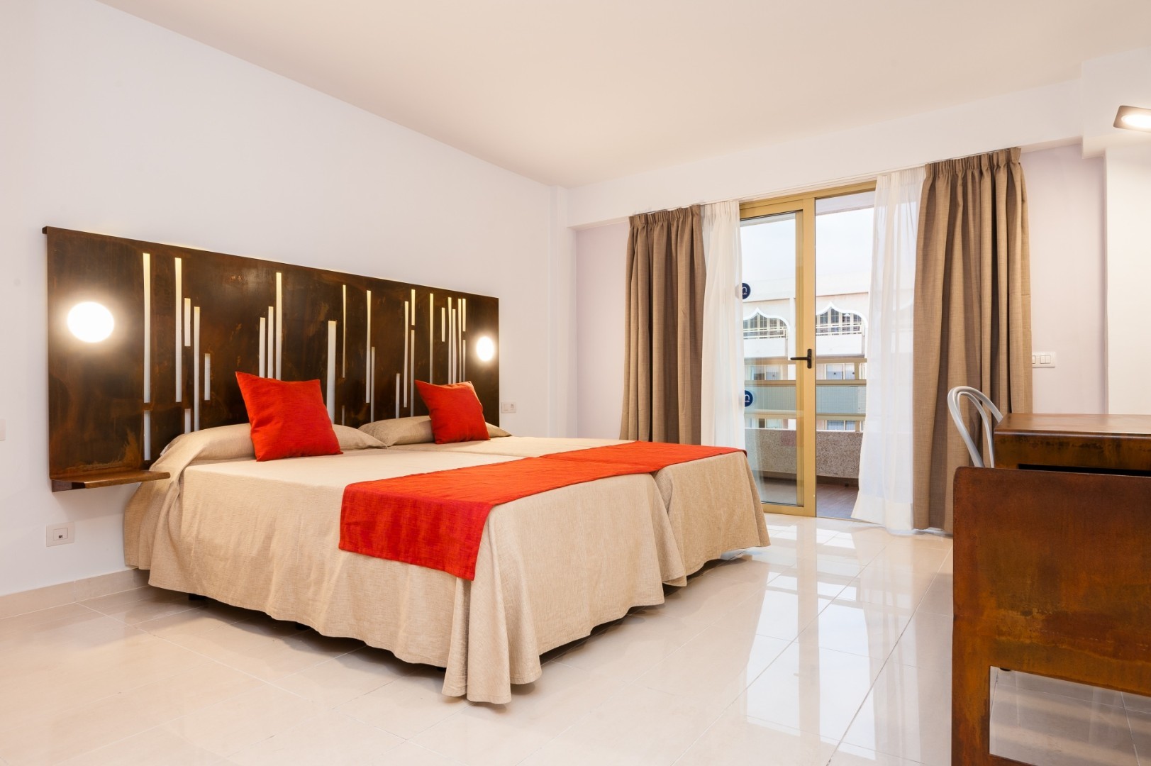 Bright twin bedroom with red accents, decorative headboard, and balcony doors with curtains.