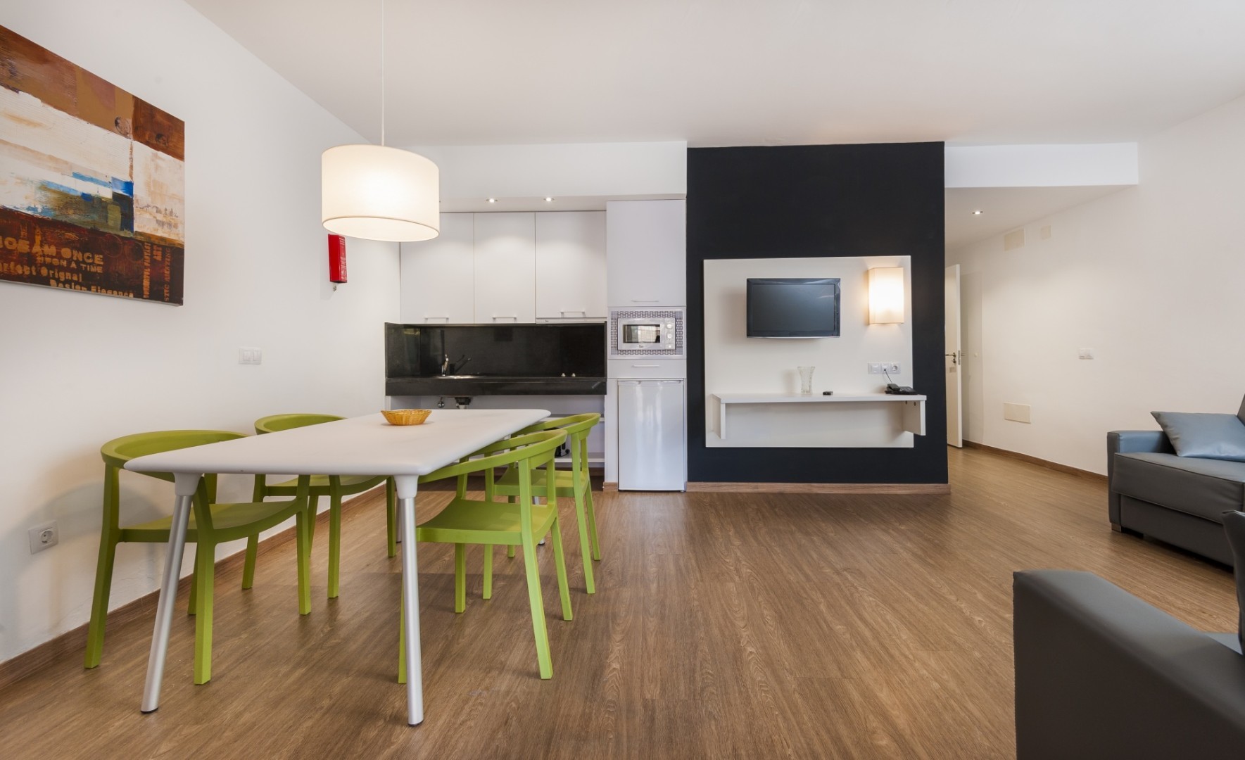 Apartment dining area with table, green chairs, kitchenette, and wall-mounted TV.