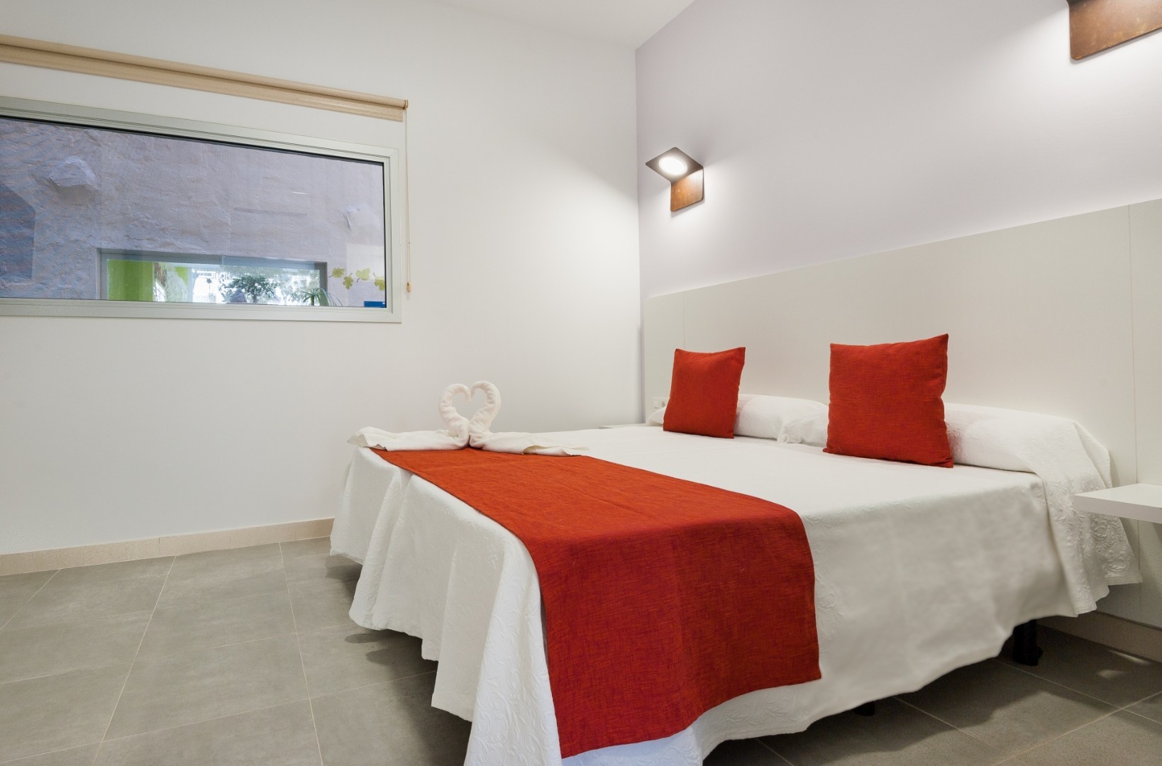 Simple double bedroom with red cushions, red bed runner, and towel-heart decoration.