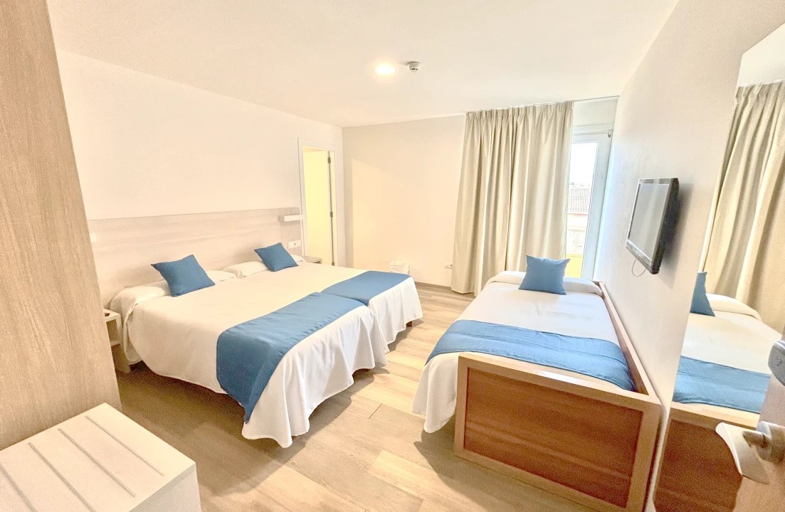 Chambre triple lumineuse avec lit double et lit simple, TV murale et rideaux.