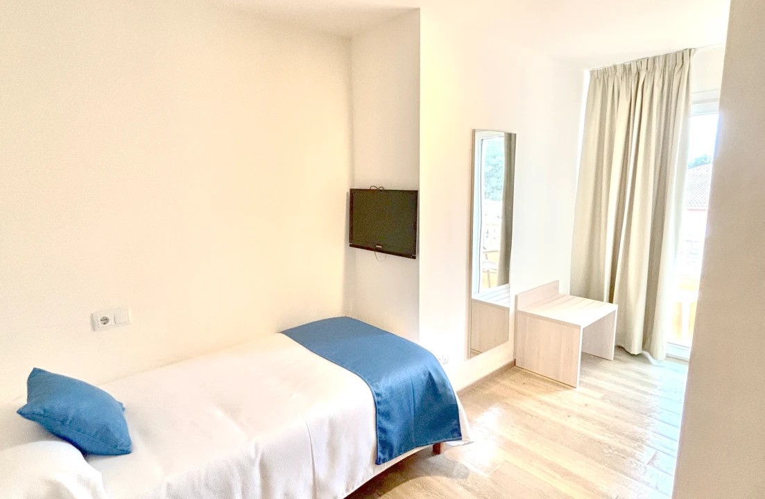 Chambre simple épurée avec bureau, miroir, TV murale et porte-fenêtre.