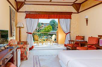 Hotel room with double bed, red decor, and balcony with garden view