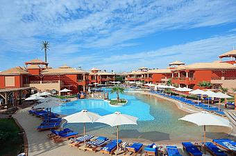 Resort pool area with red buildings, blue loungers, and umbrellas under a clear sky