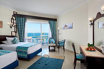 Bright hotel room with twin beds, blue decor, and balcony offering sea view, ideal for sports camp accommodation