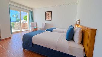 Sea-view hotel room with twin beds, blue linens, and large glass doors opening to a terrace