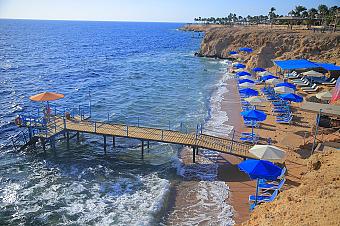 Sandy beach with blue umbrellas, sun loungers, and wooden pier extending into the clear sea
