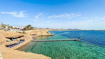 Clear turquoise bay with sunbeds, thatched umbrellas, and a wooden pier stretching into the sea