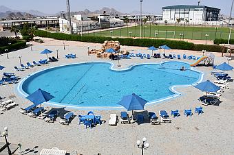 Large swimming pool surrounded by blue umbrellas and sunbeds near sports training fields