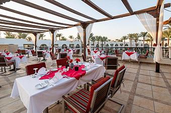 Open-air restaurant terrace with red and white table settings overlooking palm trees and resort buildings