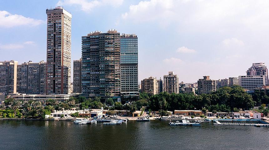 Riverfront skyline with high-rise buildings, trees, and docked boats under a partly cloudy blue sky