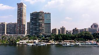 Riverfront skyline with high-rise buildings, trees, and docked boats under a partly cloudy blue sky