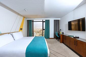 Modern hotel room with white bed and teal runner, desk and TV, large window overlooking the city