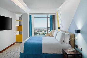 Hotel room with blue bedspread, wall TV and armchair, large window with river and city view
