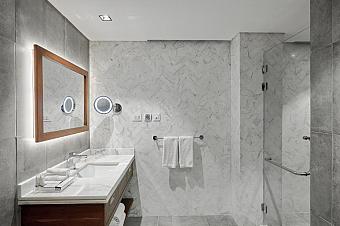 Gray-tiled hotel bathroom with marble walls, vanity sink, lit mirror, towel rack, and glass shower